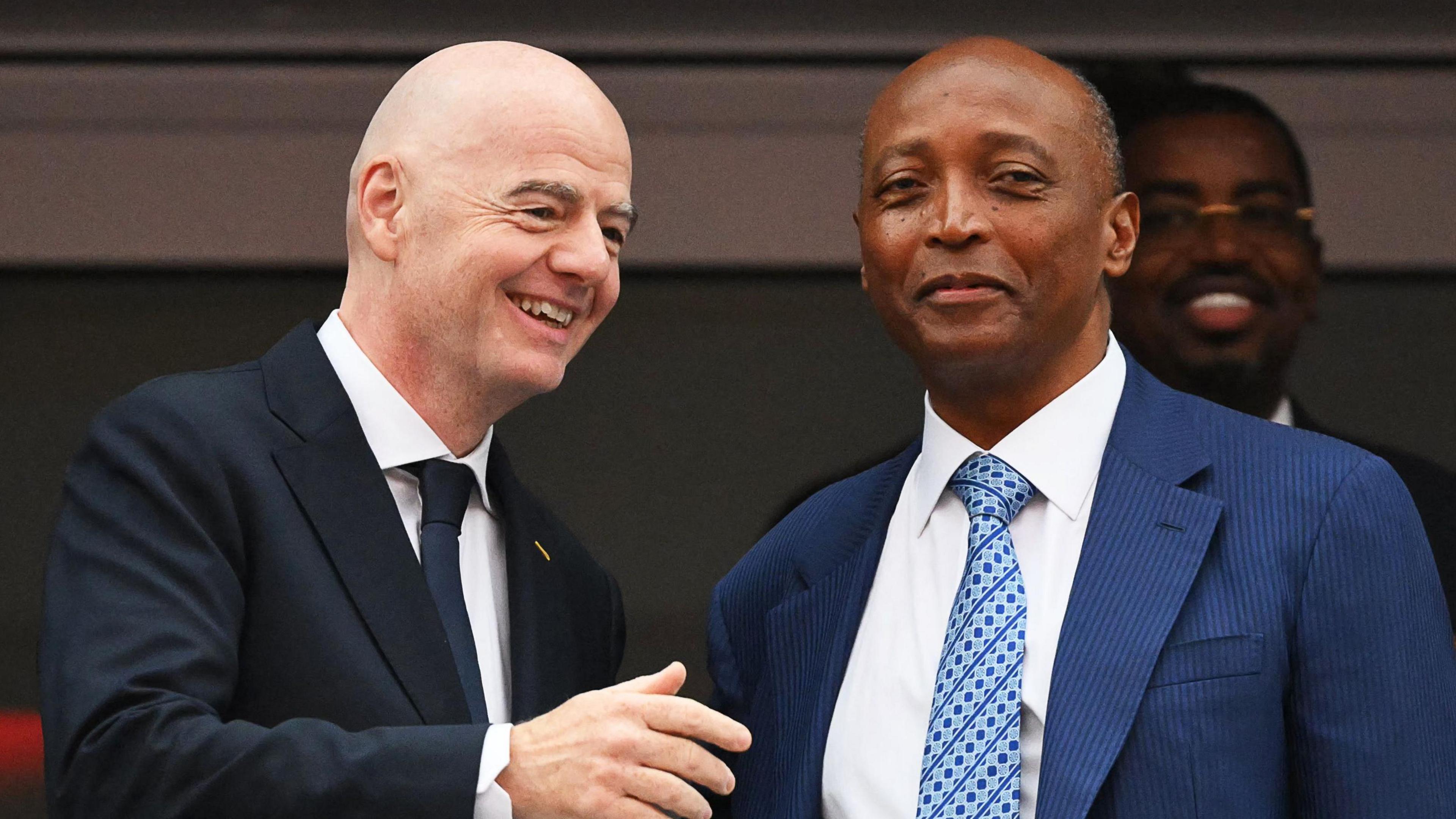 Fifa president Gianni Infantino, wearing a black jacket, white shirt and dark blue tie, smiles and holds up his right arm as he speaks to Caf president Patrice Motsepe, who is wearing a blue suit jacket, white shirt and blue patterned tie